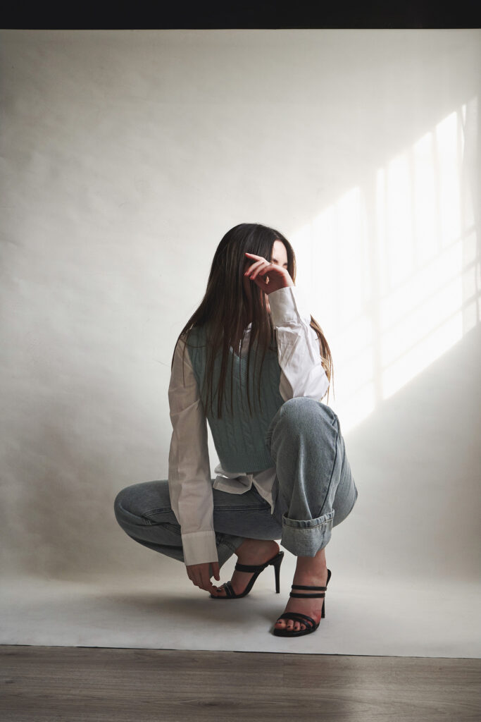 Gabriela Inzunza for Avice. A model with long hair wearing a white shirt blue vest and blue jeans poses against a white backdrop with sunlight streaming in. She's wearing black strappy heels.